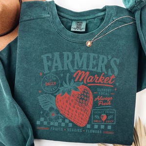 May include: A dark teal crewneck sweatshirt featuring a graphic print of a large red strawberry. The text reads Farmer's Market, Fresh Daily, Support Local, Always Fresh, Locally Grown, Sweet & Juicy, and Fruits, Veggies, Flowers in red and teal.