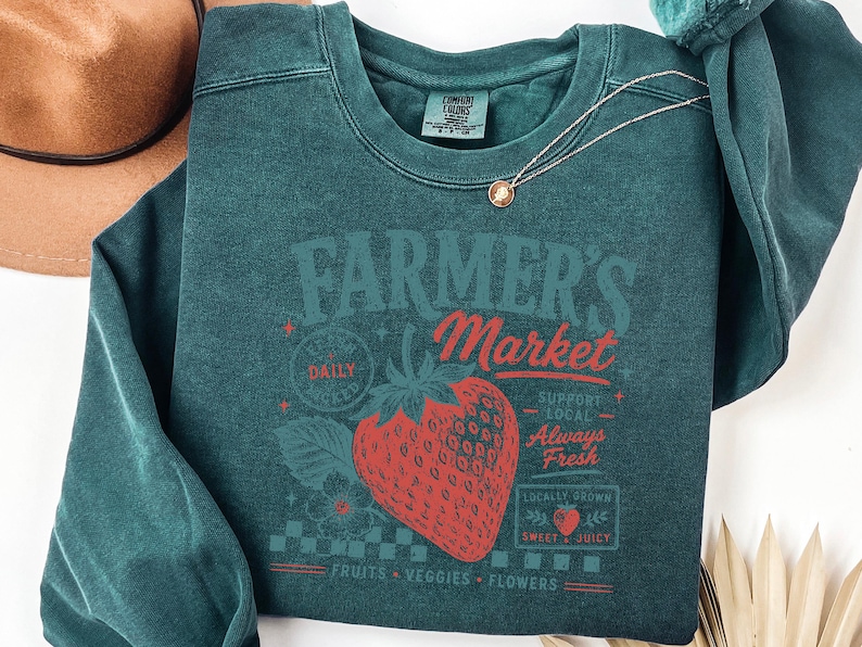 May include: A dark teal crewneck sweatshirt featuring a graphic print of a large red strawberry. The text reads Farmer's Market, Fresh Daily, Support Local, Always Fresh, Locally Grown, Sweet & Juicy, and Fruits, Veggies, Flowers in red and teal.