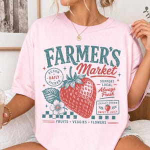 May include: A light pink t-shirt featuring a graphic print with the text Farmer's Market, Fresh Daily Picked, Support Local, Always Fresh, Locally Grown, Sweet & Juicy, and Fruits, Veggies, Flowers, centered around a large red strawberry illustration.