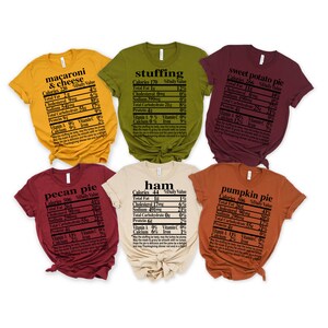 May include: A collection of six t-shirts in various colors, each featuring a nutrition facts label for Thanksgiving foods like macaroni & cheese, stuffing, and sweet potato pie. The shirts are short-sleeved.