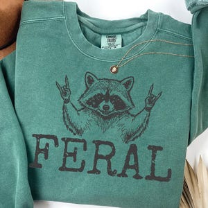 May include: A pigment-dyed green crewneck sweatshirt featuring a black graphic of a raccoon making rock-and-roll hand gestures above the word FERAL in a distressed font. The inner neck label reads COMFORT COLORS.