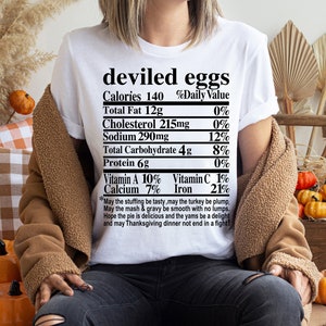 May include: White t-shirt with the words "deviled eggs" and a nutrition facts label. The shirt also includes a Thanksgiving-themed poem. The person is wearing a brown jacket and sitting near pumpkins and fall decor.