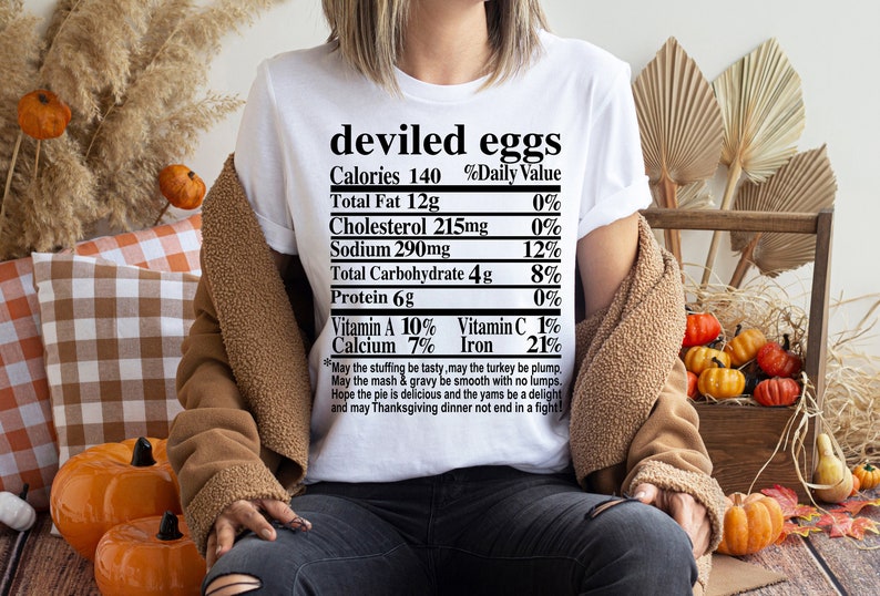May include: White t-shirt with the words "deviled eggs" and a nutrition facts label. The shirt also includes a Thanksgiving-themed poem. The person is wearing a brown jacket and sitting near pumpkins and fall decor.