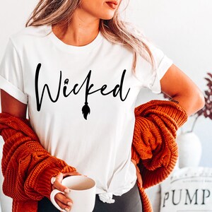 Puede incluir: Camiseta blanca con texto negro que dice "Wicked" con un fantasma y una escoba gráfica.