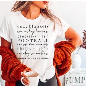Op de afbeelding: Wit T-shirt met zwarte tekst met herfstthema-zinnen: "cozy blankets", "crunchy leaves", "crackling fires", "football", "crisp mornings", "chilly nights", "comfy sweaters" en "pumpkin everything". Een oranje trui is over de schouders gedrapeerd.
