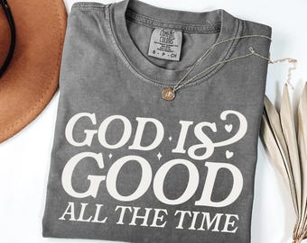 May include: A gray t-shirt with the text "GOD IS GOOD ALL THE TIME" in white script. A gold pendant necklace and a brown hat are visible. The shirt is folded, and a dried plant is on the side.