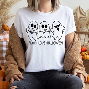 Puede incluir: Camiseta blanca con un gráfico negro de tres fantasmas y el texto "Peace Love Halloween".
