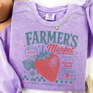 May include: A lavender crewneck sweatshirt featuring a graphic print of a large red strawberry and the text Farmer's Market, Fresh Daily Picked, Support Local, Always Fresh, Locally Grown, Sweet & Juicy, and Fruits, Veggies, Flowers in green and red.