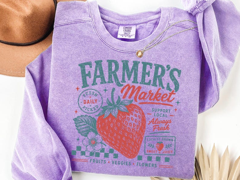 May include: A lavender crewneck sweatshirt featuring a graphic print of a large red strawberry and the text Farmer's Market, Fresh Daily Picked, Support Local, Always Fresh, Locally Grown, Sweet & Juicy, and Fruits, Veggies, Flowers in green and red.