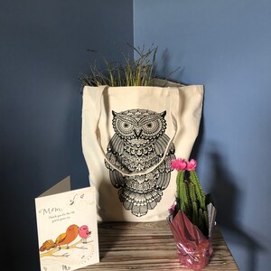 May include: A white canvas tote bag with a black owl design. The bag is filled with dried grasses. A card with the text "Mom, Thank you for the life you've given me" is on a wooden table next to a pink pot with a cactus and pink flowers.