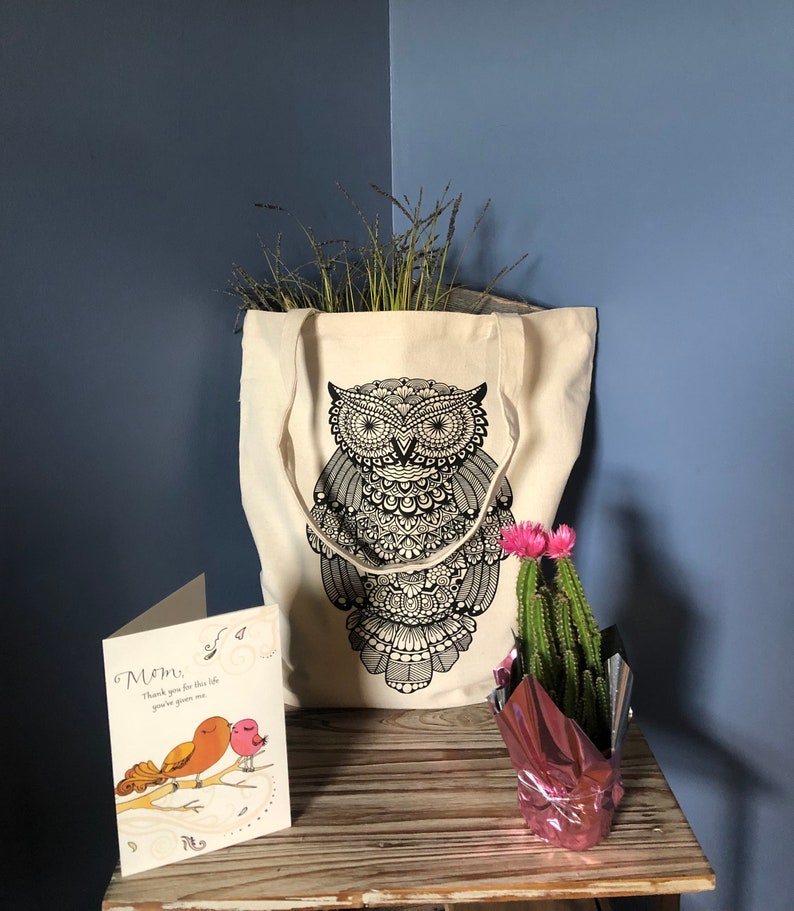 May include: A white canvas tote bag with a black owl design. The bag is filled with dried grasses. A card with the text "Mom, Thank you for the life you've given me" is on a wooden table next to a pink pot with a cactus and pink flowers.