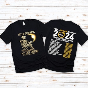 Puede incluir: Camiseta negra con un gráfico dorado de un esqueleto sentado en una silla con un eclipse solar detrás. El texto "Hello Darkness, My Old Friend - April 8th, 2024" está impreso en la camiseta. La parte posterior de la camiseta tiene un gráfico dorado con el texto "Total Solar Eclipse 2024 04.08. North America Tour 1:52 pm - 3:33 p.m." y una lista de ciudades y horarios.