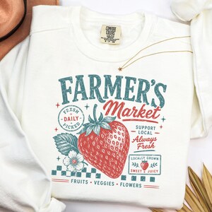 May include: A cream-colored crewneck sweatshirt featuring a graphic print with the text Farmer's Market, Fresh Daily Picked, Support Local, Always Fresh, Locally Grown, Sweet & Juicy, and Fruits Veggies Flowers, centered around a large red strawberry.