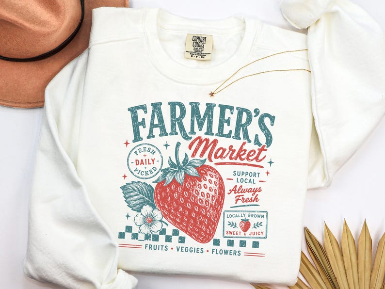 May include: A cream-colored crewneck sweatshirt featuring a graphic print with the text Farmer's Market, Fresh Daily Picked, Support Local, Always Fresh, Locally Grown, Sweet & Juicy, and Fruits Veggies Flowers, centered around a large red strawberry.