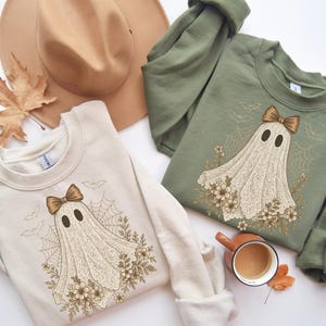 May include: Two sweatshirts, one cream and one olive green, feature a ghost design with a bow, flowers, and spiderwebs. A tan hat and a cup of coffee with a leaf are also in the image. The sweatshirts are long-sleeved.