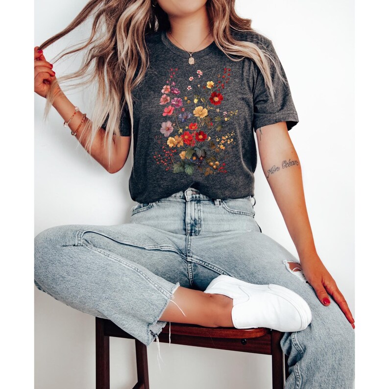 May include: A dark gray t-shirt with a floral print featuring red, yellow, pink, and purple flowers. The flowers are arranged in a bouquet shape and are surrounded by green leaves. The t-shirt is being worn by a person sitting on a stool.