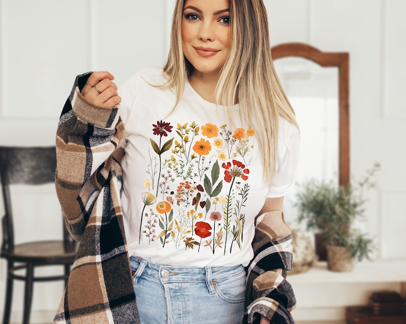 May include: A white t-shirt with a colorful floral print featuring various flowers, leaves, and stems in shades of red, orange, yellow, and green.