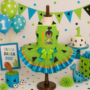 Puede incluir: Un vestido verde sin mangas con falda de tul que presenta patrones de triángulos negros, un número uno de color naranja brillante y un gráfico de personaje de dibujos animados. La cintura tiene un lazo de satén verde lima y el borde es azul.