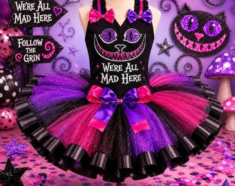 May include: A black sleeveless dress featuring a graphic of a cat face with purple eyes and a wide grin, with the text "We're All Mad Here" printed in white. The dress has a multi-layered tulle skirt in black, purple, and pink with ribbon bows.