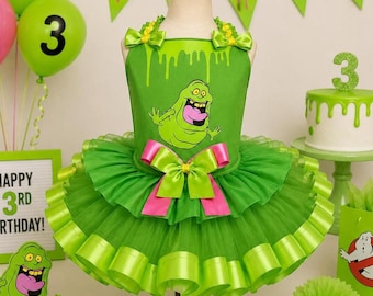 May include: A bright green dress featuring a graphic of a cartoon ghost, a layered tulle skirt with ribbon trim, and a large pink and green bow at the waist. The bodice has a slime-drip pattern at the top and small bows on the shoulder straps.