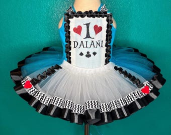 May include: A blue and white tutu dress with a black and white zigzag trim. The bodice has a white rectangle with the text "I DALANI" and red heart and black suit symbols. The tutu has layers of blue and white tulle.