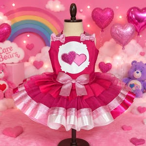 May include: A dark pink dress featuring a white circular patch with two hearts on the chest, small bows on the shoulder straps, and a large shimmering bow at the waist. The skirt is made of layered tulle with a wide, metallic pink ribbon trim.