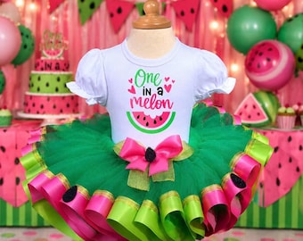 以下が含まれることがあります： One in a melonの文字とスイカのグラフィックがプリントされた白い半袖シャツと、鮮やかなピンクのリボンで縁取られた緑色の重ね着チュールチュチュスカート。ウエストには黒い種のようなアクセントが付いた大きなピンクのリボンがあしらわれています。