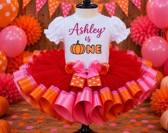 May include: A white short-sleeved bodysuit with ruffled sleeves featuring the text Ashley is one and a pumpkin graphic in orange and pink. The bodysuit is attached to a multi-layered red, orange, and pink tulle tutu skirt with a large bow.