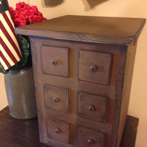 May include: A brown wooden cabinet with six square drawers and metal knobs. The cabinet has a slightly distressed finish with blue accents. An American flag and red flowers are visible in the background.