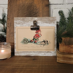May include: A whitewashed wood picture frame with a metal clip holding a vintage Christmas card. The card features a child pulling a Christmas tree with a small dog following behind. The text on the card reads "I'm wishing a happy Christmas for a happy child I know."