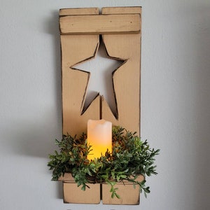 May include: A rustic wooden candle holder with a star cutout and a small shelf. The holder is painted a light yellow color and has a distressed finish. A green wreath surrounds a candle on the shelf. The text "Barn Door Primitives" is printed below the holder.