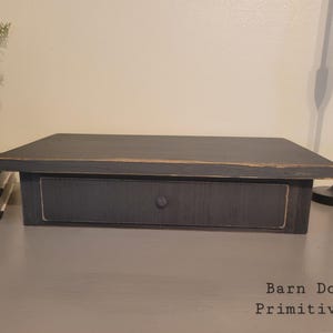 May include: A black, rectangular wooden riser with a drawer, featuring a knob. The distressed finish gives it a rustic, farmhouse aesthetic. The text "Barn Door Primitives" is visible.