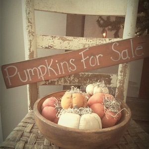 May include: A wooden bowl filled with fabric pumpkins in shades of orange, white, and brown. The bowl is sitting on a woven chair seat. A sign above the bowl reads "Pumpkins For Sale".
