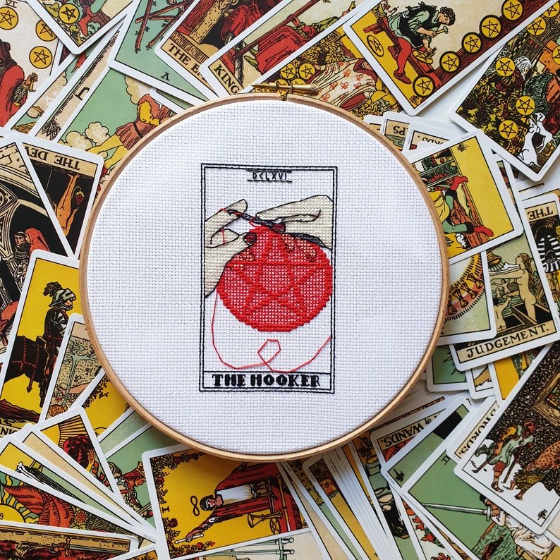 May include: A white cross-stitch embroidery hoop with a red and white design of a hand holding a ball of yarn. The text "The Hooker" is printed below the design.
