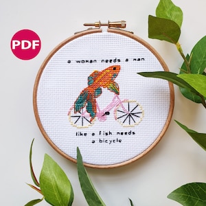 May include: A white embroidery hoop with a cross-stitch design of a goldfish riding a bicycle. The text "a woman needs a man like a fish needs a bicycle" is stitched in black thread.