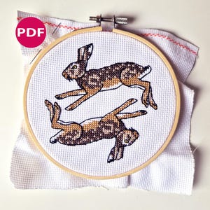 May include: A cross-stitch embroidery featuring two brown and tan hares in mid-leap, set against a white fabric background. The design is framed within a wooden embroidery hoop. A pink circle with the letters "PDF" is in the upper left corner.