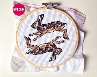 May include: A cross-stitch embroidery featuring two brown and tan hares in mid-leap, set against a white fabric background. The design is framed within a wooden embroidery hoop. A pink circle with the letters "PDF" is in the upper left corner.