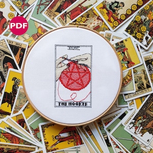 Funny tarot-inspired cross stitch pattern PDF titled 'The Hooker' in bold black text with pink details, shown framed in embroidery hoop. The image shows a pair of hands crocheting a round pentagram, framed in the style of a tarot card. The image also shows tarot cards strewn in the background - these are not included in the listing, and only serve for decoration.