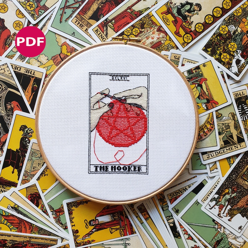 Funny tarot-inspired cross stitch pattern PDF titled 'The Hooker' in bold black text with pink details, shown framed in embroidery hoop. The image shows a pair of hands crocheting a round pentagram, framed in the style of a tarot card. The image also shows tarot cards strewn in the background - these are not included in the listing, and only serve for decoration.