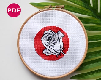 May include: A white rose cross stitch pattern with a red circle background. The pattern is in a wooden embroidery hoop.