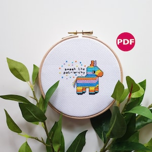 May include: A cross-stitch pattern featuring a colorful pinata with the text "Smash the patriarchy" in a white circle with yellow stars. The pattern is in a wooden embroidery hoop.