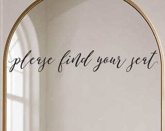 May include: An arched mirror with a thin gold frame featuring the black cursive text "please find your seat" applied to the glass surface. The mirror reflects a bright interior space and is positioned next to white floral stems.
