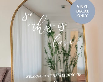 May include: A gold-framed arched mirror with a white vinyl decal that reads "So this is love" and "Welcome to the wedding of Lisa and Aaron June 28th, 2024".