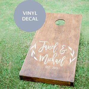 May include: A wooden cornhole board with a hole at the top and the text "Janel & Michael EST. 2019" in white script. The words "VINYL DECAL" are in a light blue circle in the upper left corner. The board is on green grass.