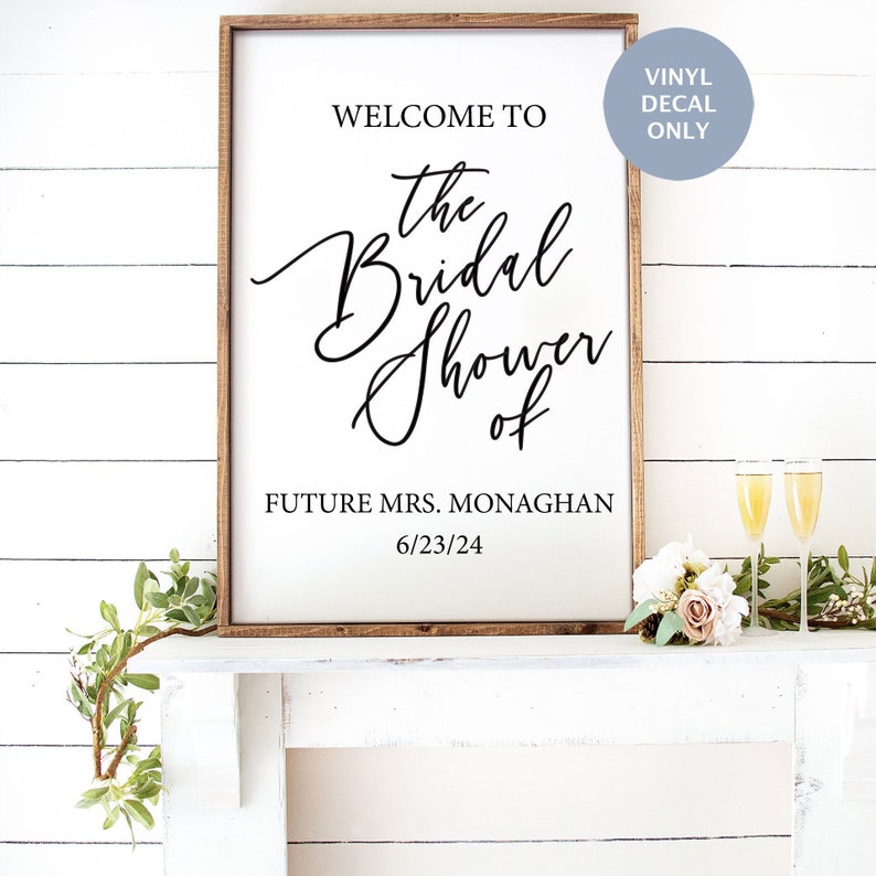 May include: A white sign with a brown wooden frame, announcing "Welcome to the Bridal Shower of Future Mrs. Monaghan 6/23/24". The sign is on a white mantel with flowers and champagne glasses.