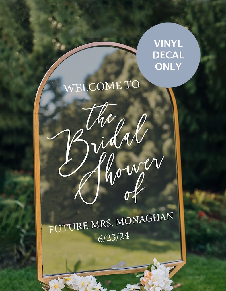 May include: A gold-framed mirror with a white vinyl decal that reads "Welcome to the Bridal Shower of Future Mrs. Monaghan 6/23/24". The decal is on a clear mirror surface. The mirror is in front of a green bush.