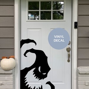 Op de afbeelding: Een witte voordeur met een zwarte vinyl sticker van een cartoon monster met scherpe tanden. De sticker zit aan de rechterkant van de deur. De deur heeft een glazen raam met een Ring deurbel. De tekst "VINYL DECAL" staat op een blauwe cirkel.