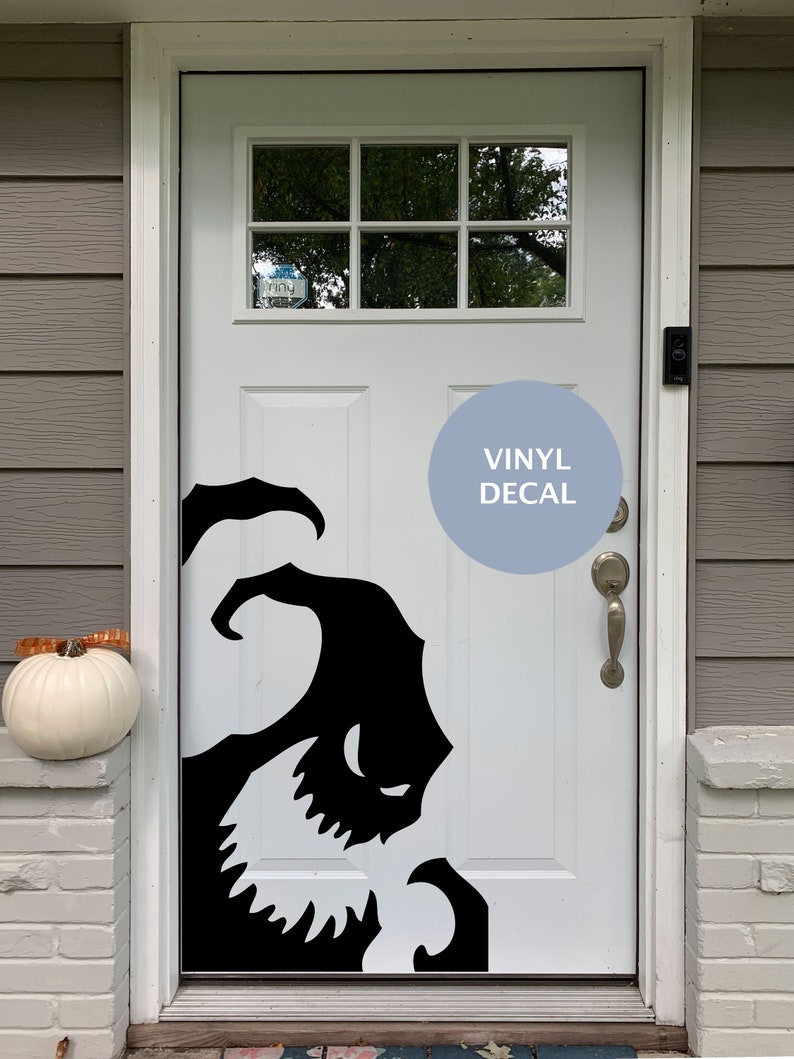 Op de afbeelding: Een witte voordeur met een zwarte vinyl sticker van een cartoon monster met scherpe tanden. De sticker zit aan de rechterkant van de deur. De deur heeft een glazen raam met een Ring deurbel. De tekst "VINYL DECAL" staat op een blauwe cirkel.