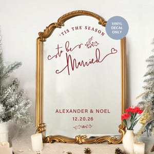 Puede incluir: Espejo dorado con marco ornamentado. El espejo muestra el texto burdeos « 'Tis the Season to be Married », los nombres « Alexander & Noel » y la fecha « 12.20.26 ». Una etiqueta de vinilo es visible.
