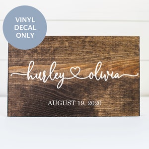 May include: A wooden sign with the names "Hurley & Olivia" and the date "August 19, 2020" written in white vinyl lettering. The sign is a dark brown color and has a rustic look.